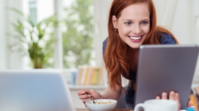 Frau sitzt am Tisch und eröffnet ein Tagesgeldkonto online Frau isst Müsli und schaut dabei strahlend auf ihren Computer Monitor