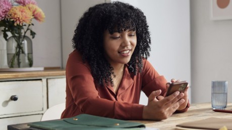 Frau sitzt am Küchentisch und sieht sich auf ihrem Handy die Zinsen für ein Tagesgeldkonto an. Frau sitzt am Küchentisch und sieht sich auf ihrem Handy die Zinsen für ein Tagesgeldkonto an.
